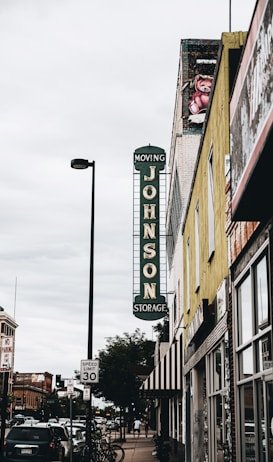 About Us Augusta GA 3 A street view featuring a vintage vertical sign that reads 'Johnson Storage Moving' on the side of a building. The surrounding area includes various storefronts, parked cars along the sidewalk, and a speed limit sign indicating 30 mph. A painted teddy bear mural can be seen near the top of one building, and the sky is overcast, adding a muted tone to the scene.